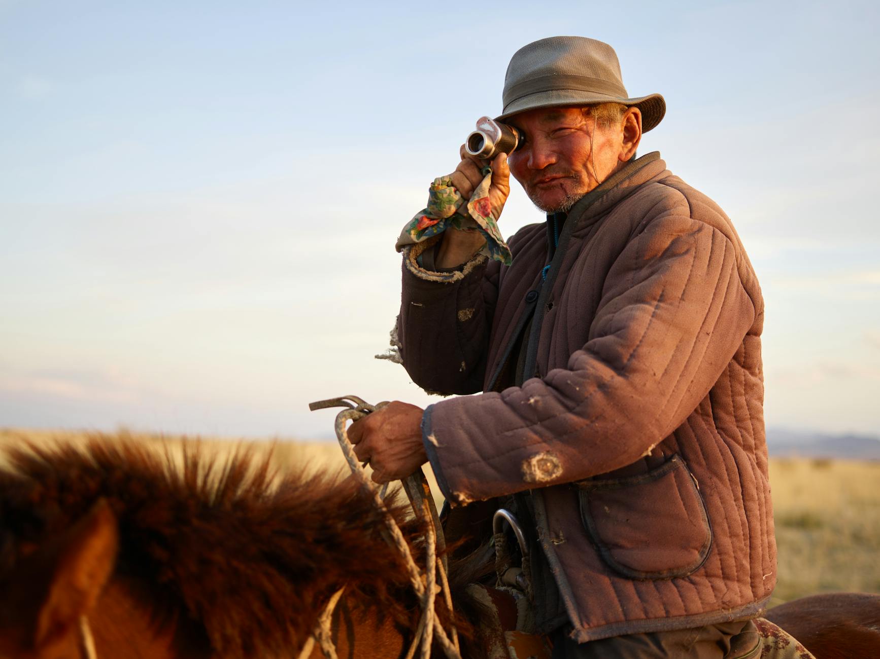 A Mongolian herder on horseback in traditional attire, using a telescope to survey the vast steppe, epitomizing the enduring nomadic culture and connection to the land.