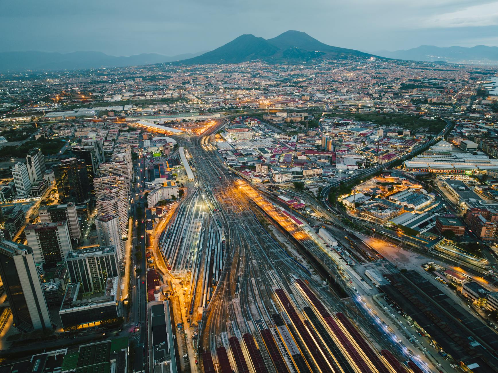 Aerial view of Naples at dusk, showcasing the vibrant cityscape with Mount Vesuvius in the background, reflecting the dynamic urban life of the Neapolitan people.
