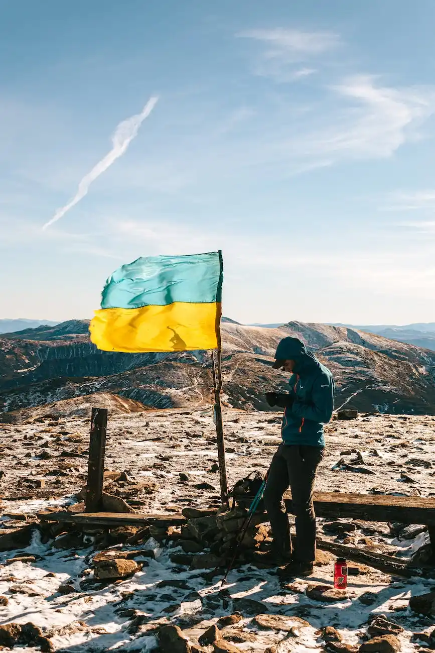 A hiker on a mountain summit beside the Ukrainian flag uses a mobile phone for Ukrainian to English translation, embodying the connection of languages across vast landscapes.