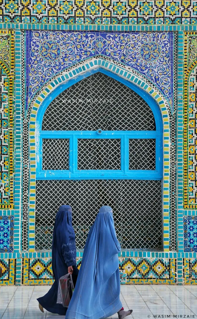 Two women in traditional blue attire walking past an intricately designed mosaic wall with geometric and floral patterns in a blend of blues, yellows, and greens, reflecting the rich cultural tapestry relevant to Dari translation.