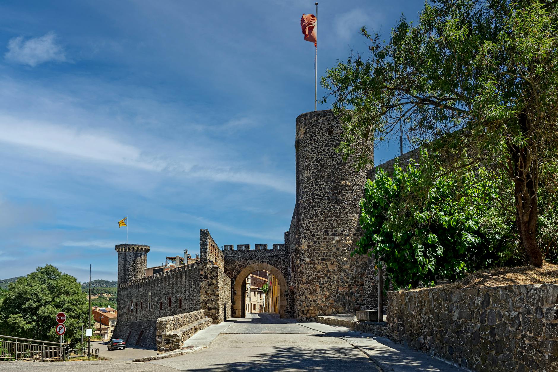 A medieval stone castle gate with tall towers and battlements, located in a picturesque town, showcasing the historical architecture of Catalonia under a bright blue sky with lush greenery around.
