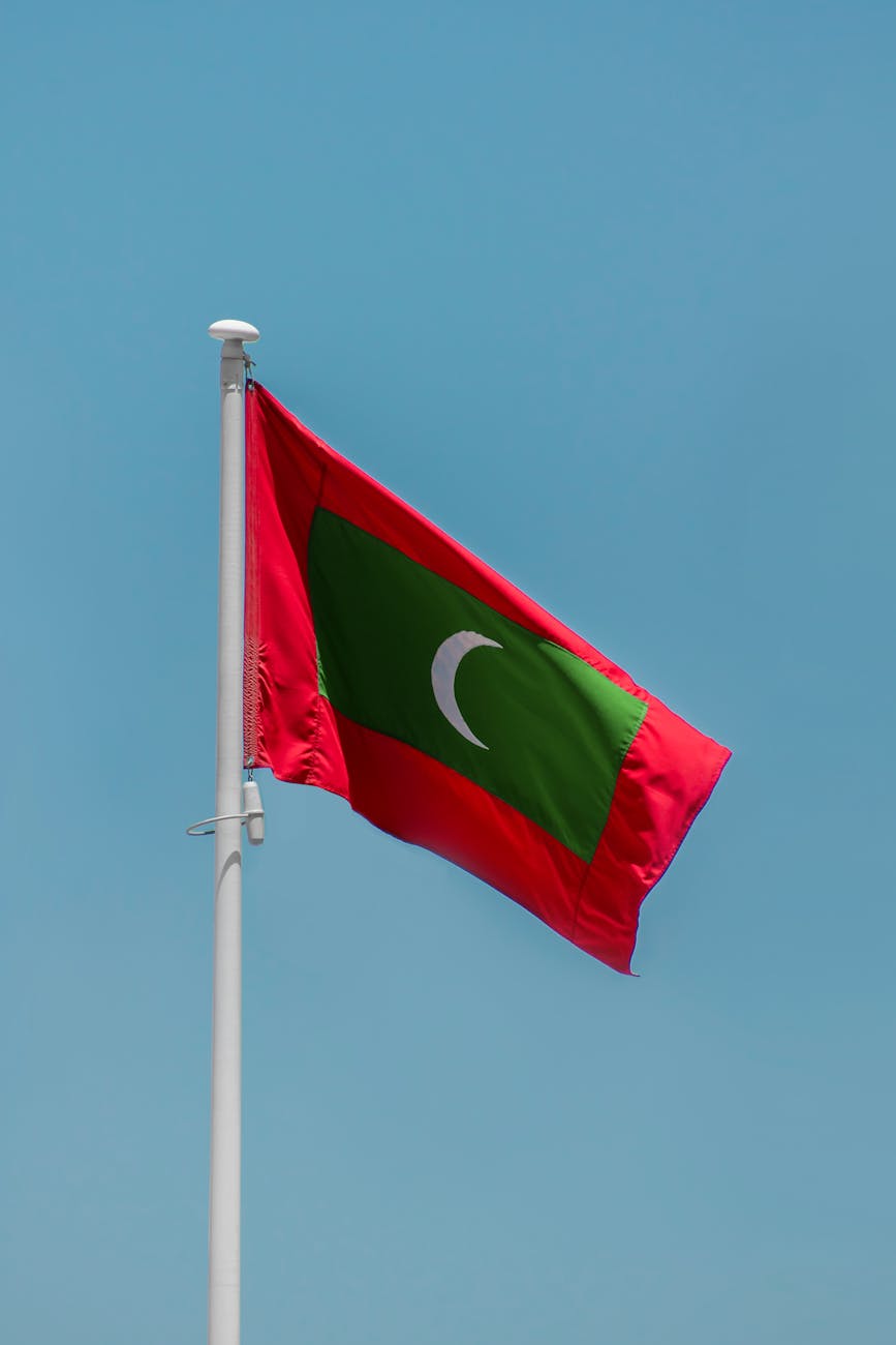 The national flag of the Maldives, featuring a red field with a green rectangle and a white crescent moon, waving against a clear blue sky.