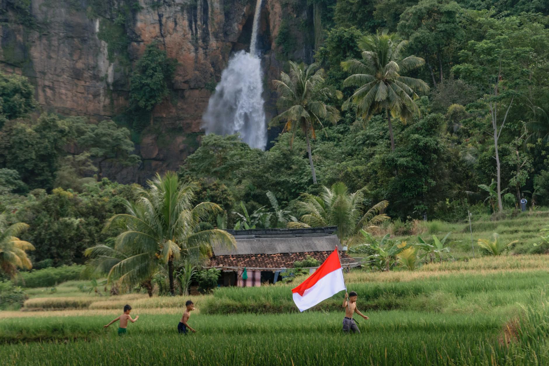 Children running through a lush green field in Aceh, Indonesia, with a waterfall in the background, carrying the Indonesian flag, symbolizing the vibrant heritage and natural beauty of Aceh.