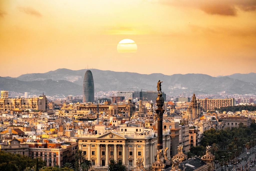 A panoramic view of a sunset over a historic cityscape, featuring a prominent statue of Christopher Columbus, iconic modern architecture, and the mountains in the background.