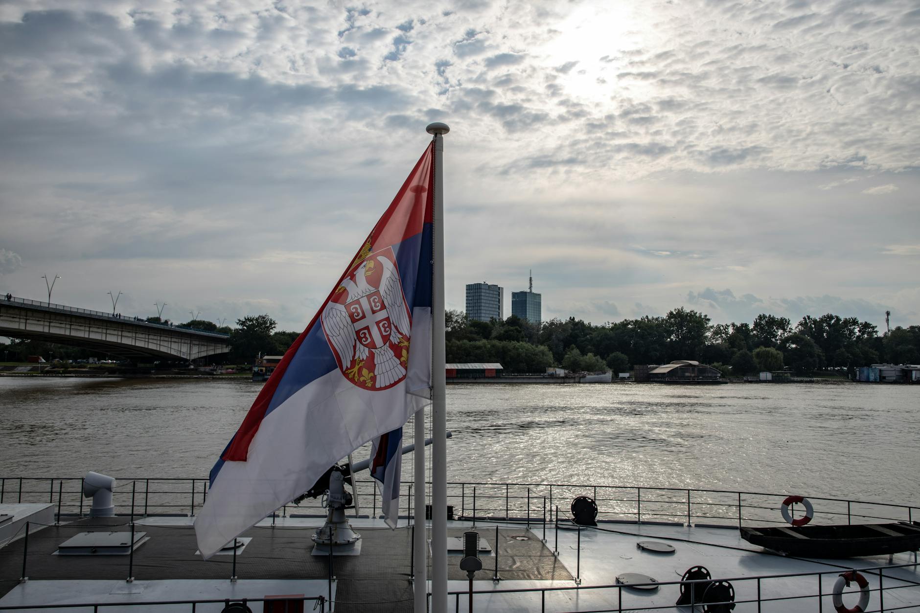 A Serbian flag fluttering on a boat, with the river and cityscape of Belgrade in the background under a cloudy sky.