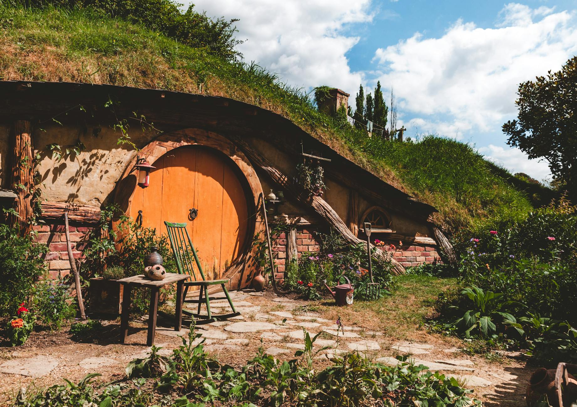 A charming hobbit-style house with a round wooden door set into a grassy hillside, surrounded by lush greenery and a clear blue sky.