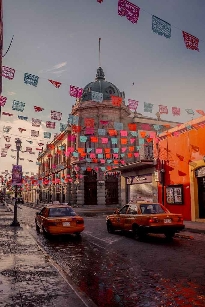 A vibrant street in Oaxaca, Mexico, adorned with colorful papel picado banners, with historic colonial buildings and traditional taxis adding to the lively atmosphere.