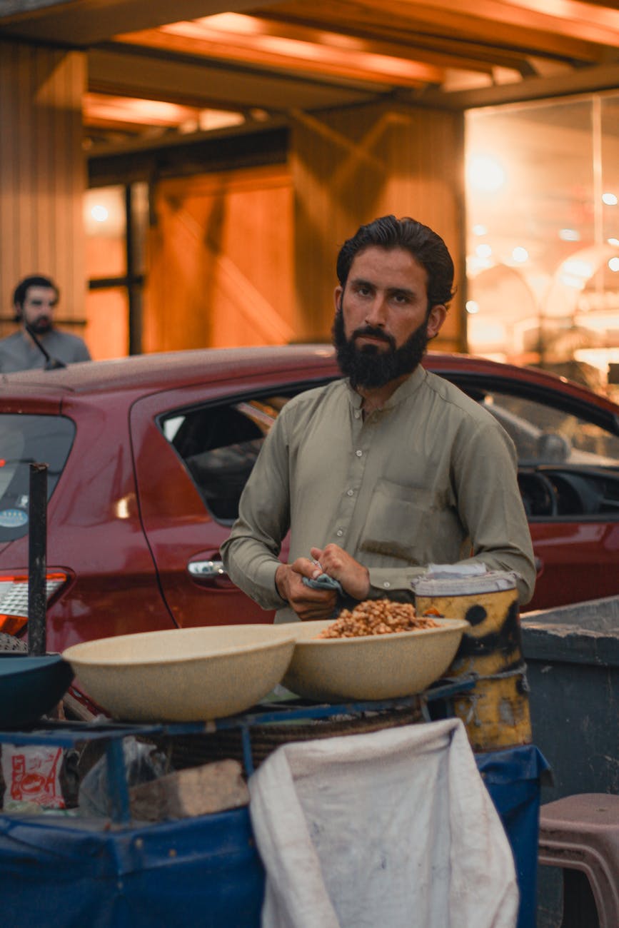 A Baloch man in traditional attire standing by a roadside market stall, symbolizing the rich cultural heritage of the Balochi tribe.
