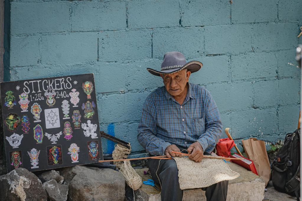 A man in traditional attire weaving near a display of colorful stickers, reflecting the cultural richness of the Sipakapense community in Guatemala.