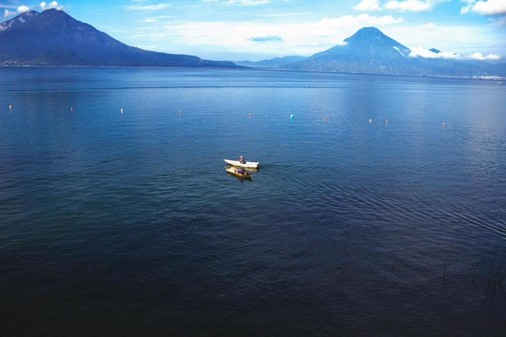 A lone boat floats on the tranquil waters of Lake Atitlán, with the majestic volcanic peaks of Guatemala rising in the background under a clear blue sky.