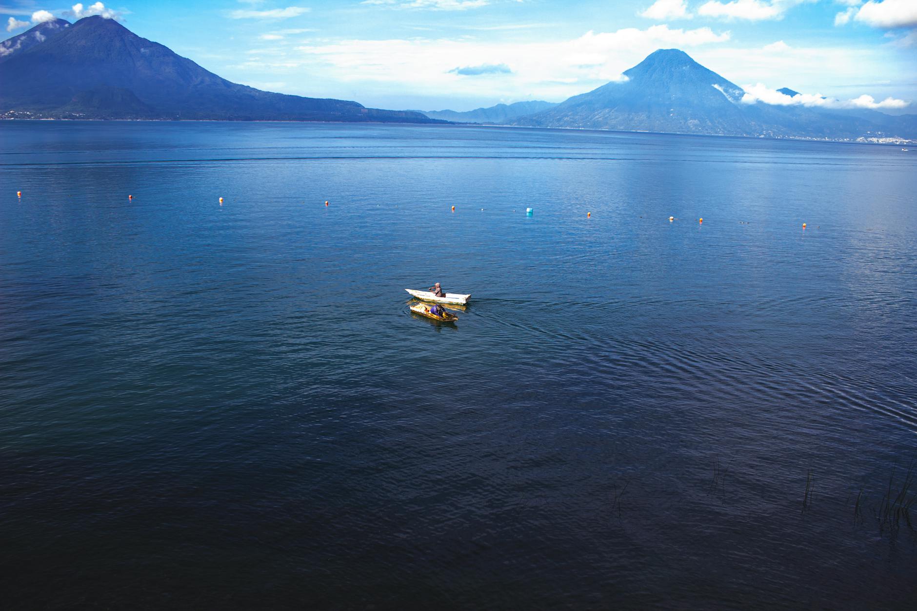 A lone boat floats on the tranquil waters of Lake Atitlán, with the majestic volcanic peaks of Guatemala rising in the background under a clear blue sky.