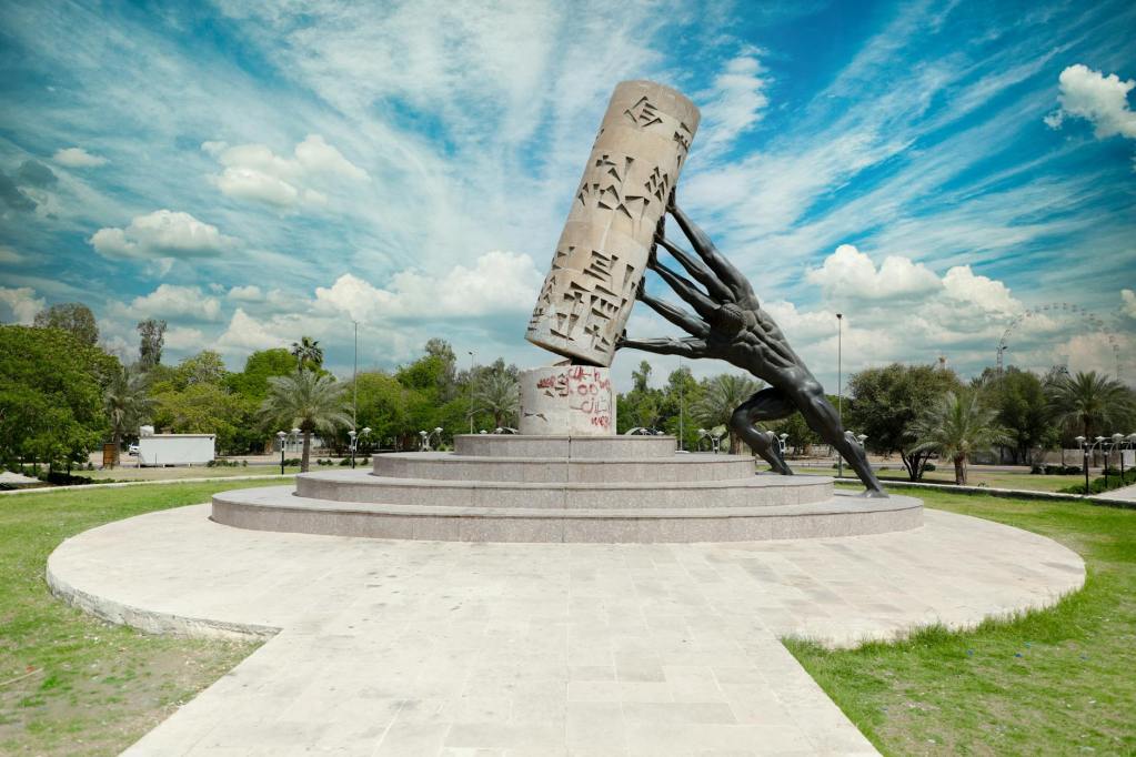 Monument in a public park symbolizing cultural strength and unity, with a large pillar being lifted by multiple figures. The pillar features inscriptions, representing the significance of preserving and translating languages like Kurdish Sorani.