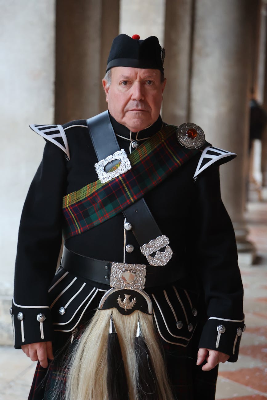 A man in traditional Scottish attire, wearing a kilt and a formal jacket, standing in front of stone columns.
