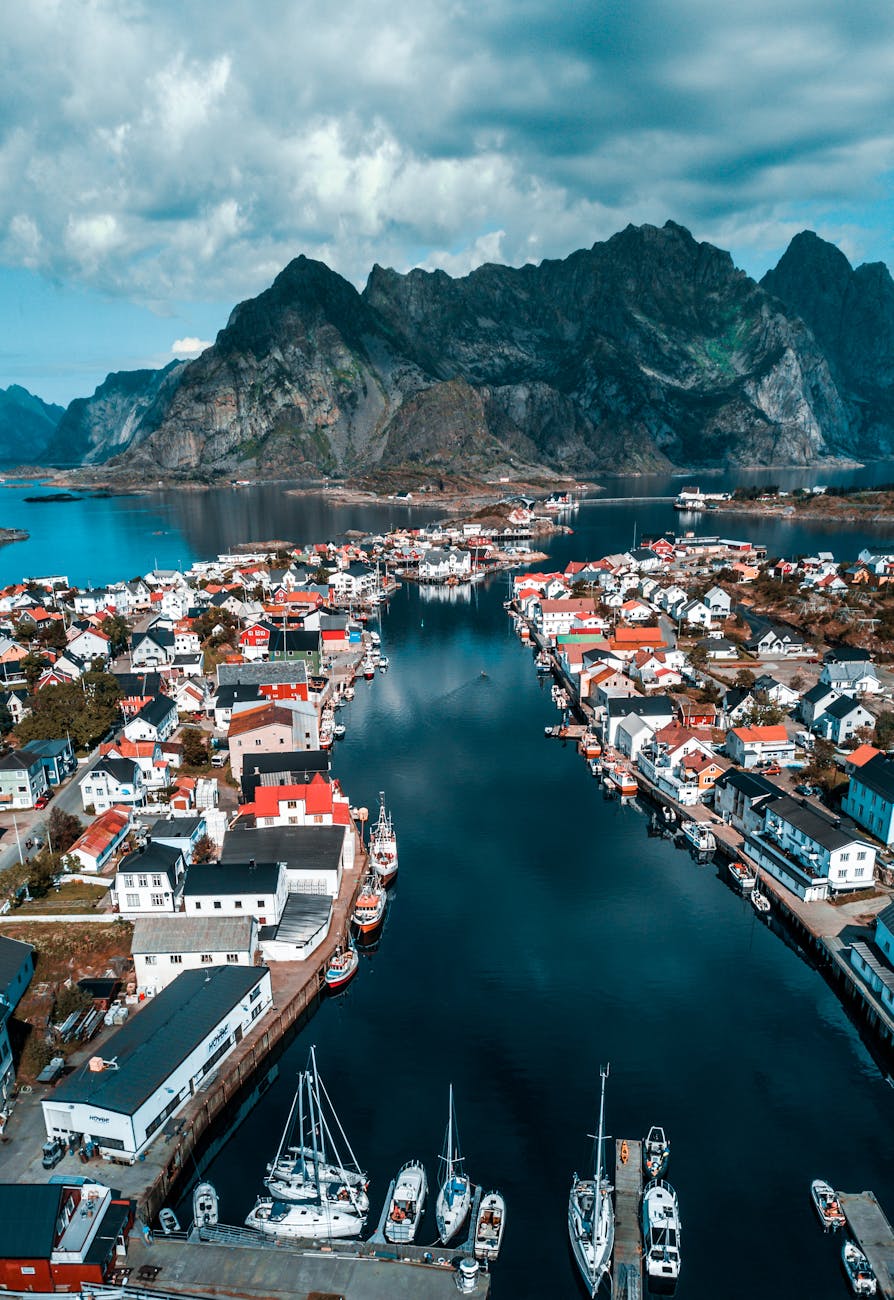 Aerial view of a picturesque coastal village with colorful houses and boats, set against a backdrop of majestic mountains under a cloudy sky.