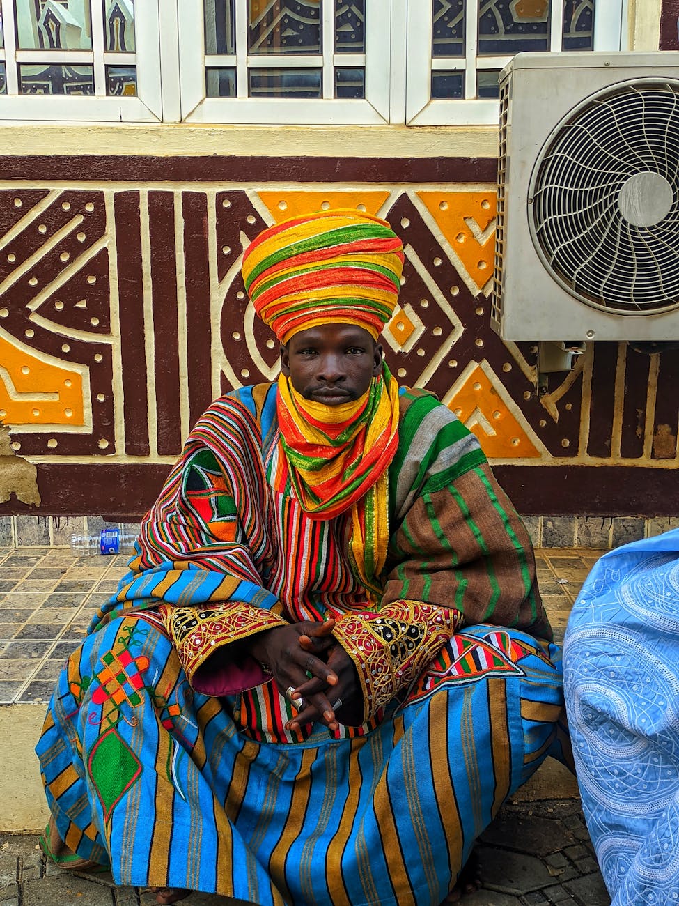 A person wearing colorful traditional West African attire, sitting in front of a patterned wall.
