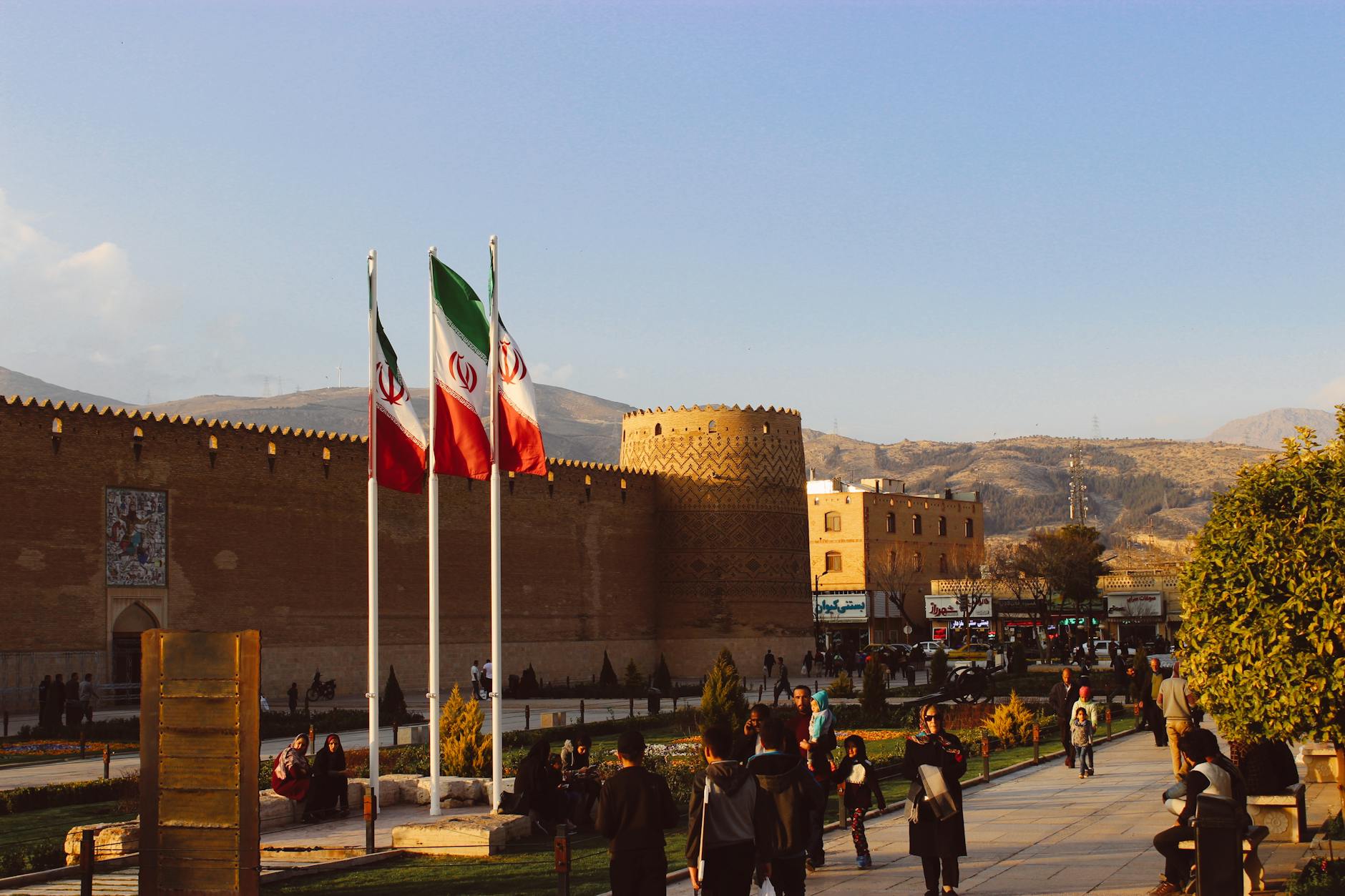 A scenic view of a historical fortress in Iran, with three Iranian flags waving in the foreground and people walking in the public square, reflecting Persian culture and heritage.